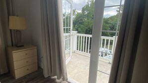 A room with a wooden floor, a bedside table with a lamp, and a view of a balcony with a railing and greenery outside. - Amazing Property Close To Beaches/Golf Course/Railway Trails (Warwick)