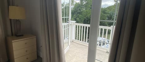 Une pièce avec un plancher en bois, une table de chevet avec une lampe, et une vue sur un balcon doté d’une balustrade et de plantes à l’extérieur.