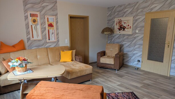 Un salon comprenant un canapé, un fauteuil, une table basse et un tapis. Les murs sont ornés de tableaux encadrés, et une porte avec un panneau de verre est visible.