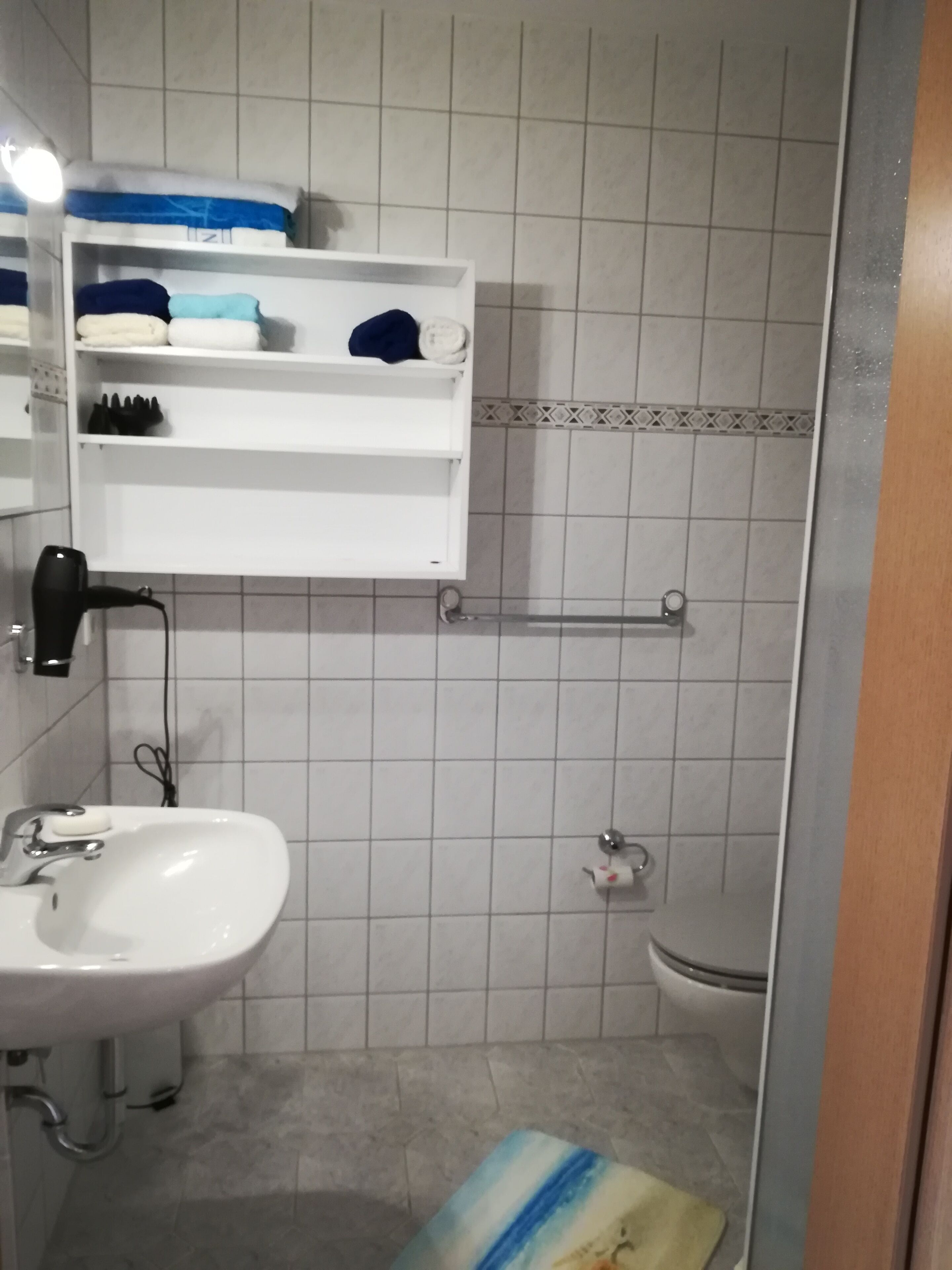 Ett badrum med ett vitt handfat, en toalett och en duschhörna. Ovanför handfatet finns en hylla med handdukar och toalettartiklar.