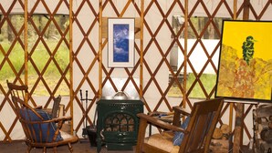 Une pièce rustique aux murs recouverts de treillis en bois, avec un poêle vert, deux chaises en bois et un tableau accroché au mur.