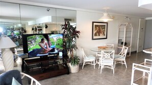 A modern living room with a flat-screen TV, white wicker furniture, a dining area, and a large potted plant. - Oceanfront, adjacent to North Ka'anapali Beach, Central A/C! Newly reduced rates (Lahaina)