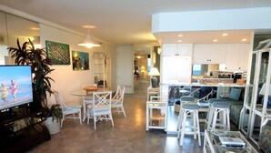A modern living room with a flat-screen TV, white wicker furniture, a glass coffee table, and a kitchen area with white cabinets and a dining table. - Oceanfront, adjacent to North Ka'anapali Beach, Central A/C! Newly reduced rates (Lahaina)