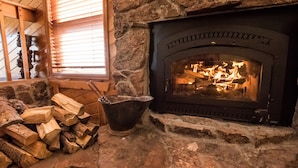 Une cheminée en pierre avec un feu qui crépite à l’intérieur, une fenêtre avec des stores et une pile de bois de chauffage.