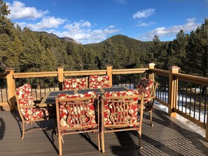 A deck with a table and chairs, overlooking a snowy landscape and forested mountains.