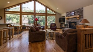 A spacious living room with large windows, a stone fireplace, wooden furniture, and a dining area.