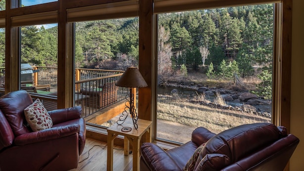 A cabin with a large window overlooking a forested area. Inside, there is a leather sofa and a small table with a lamp.