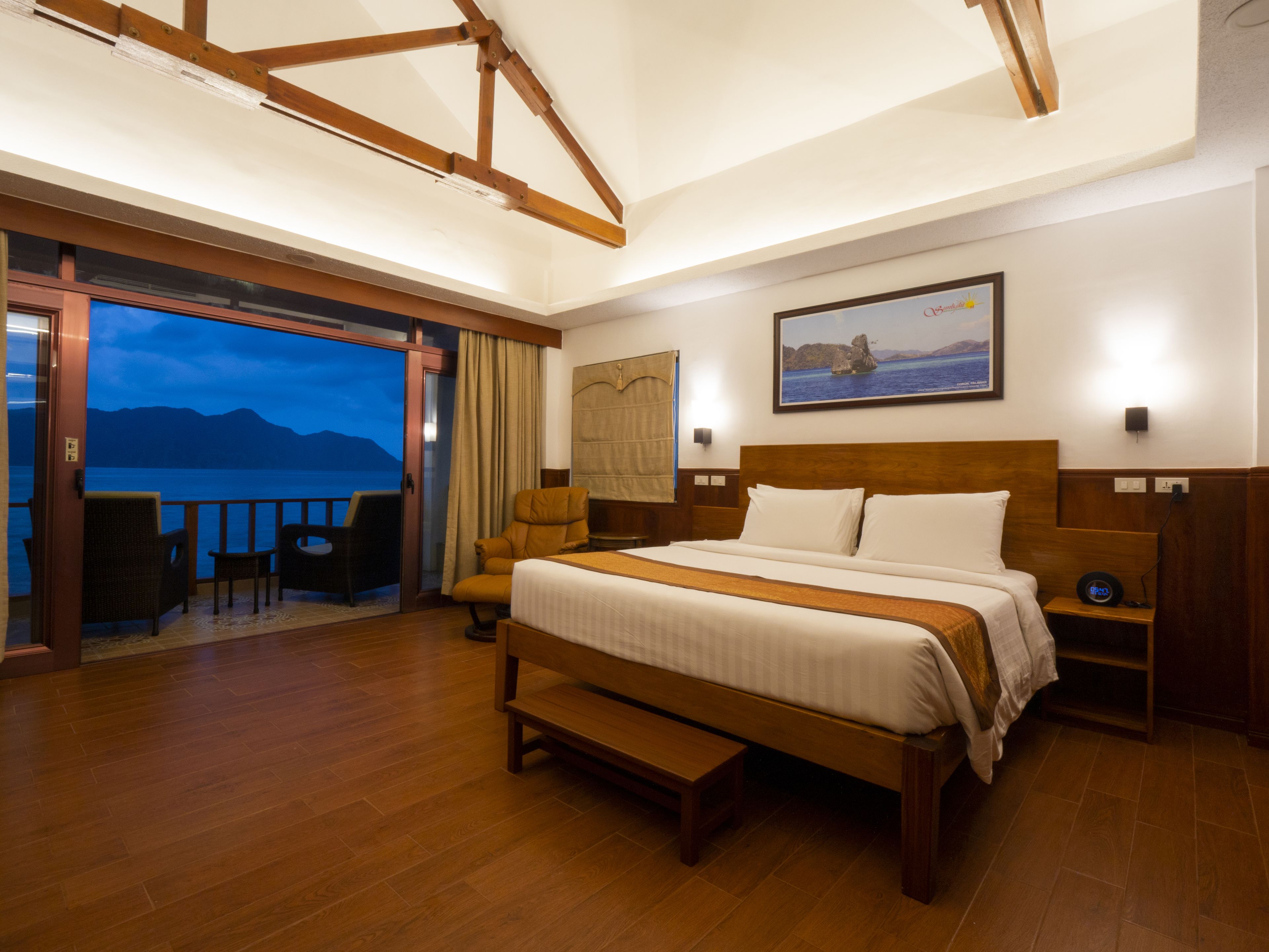 Habitación de hotel con una cama grande, una banca, suelo de madera, vista al mar y un balcón con sillas.