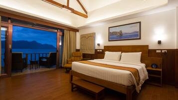 Habitación de hotel con una cama grande, una banca, suelo de madera, vista al mar y un balcón con sillas.