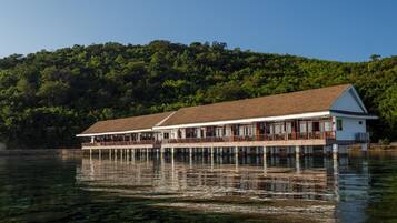 A waterfront building with a long, covered deck and multiple levels, surrounded by clear water and a forested hillside.