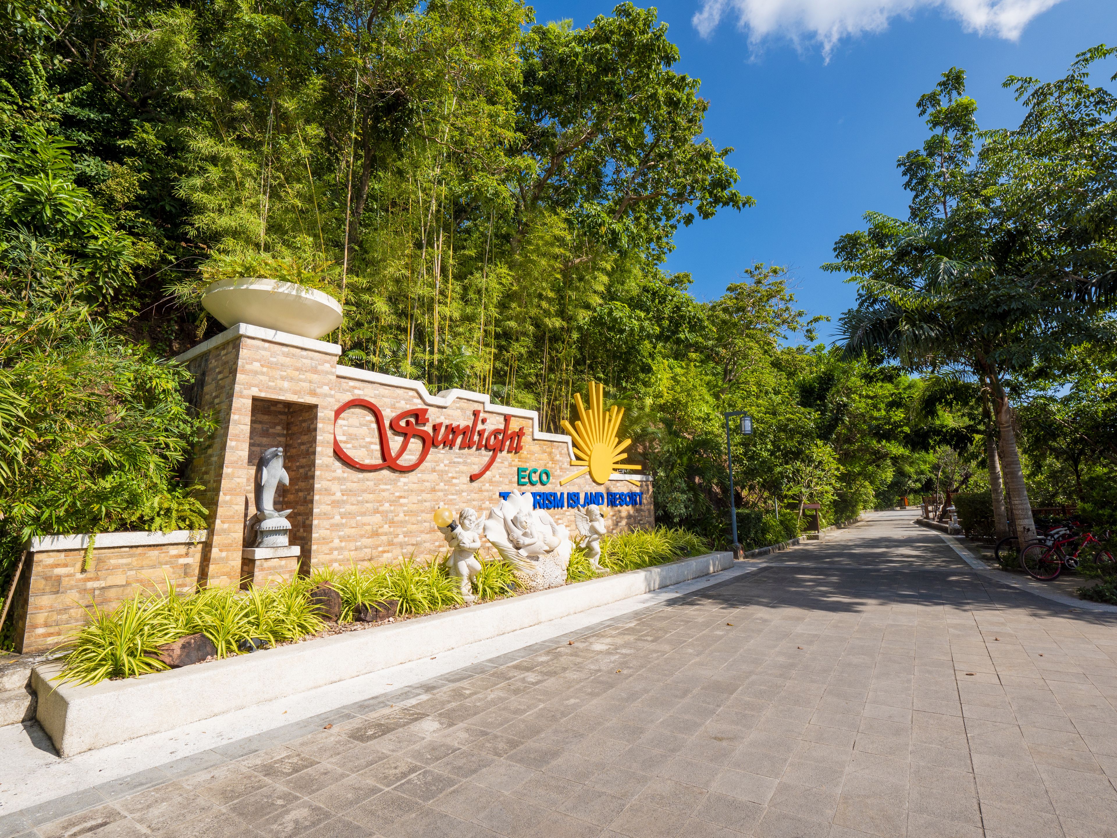 A sign for Sunlight Eco Resort with a sun emblem, surrounded by lush greenery and a paved pathway.