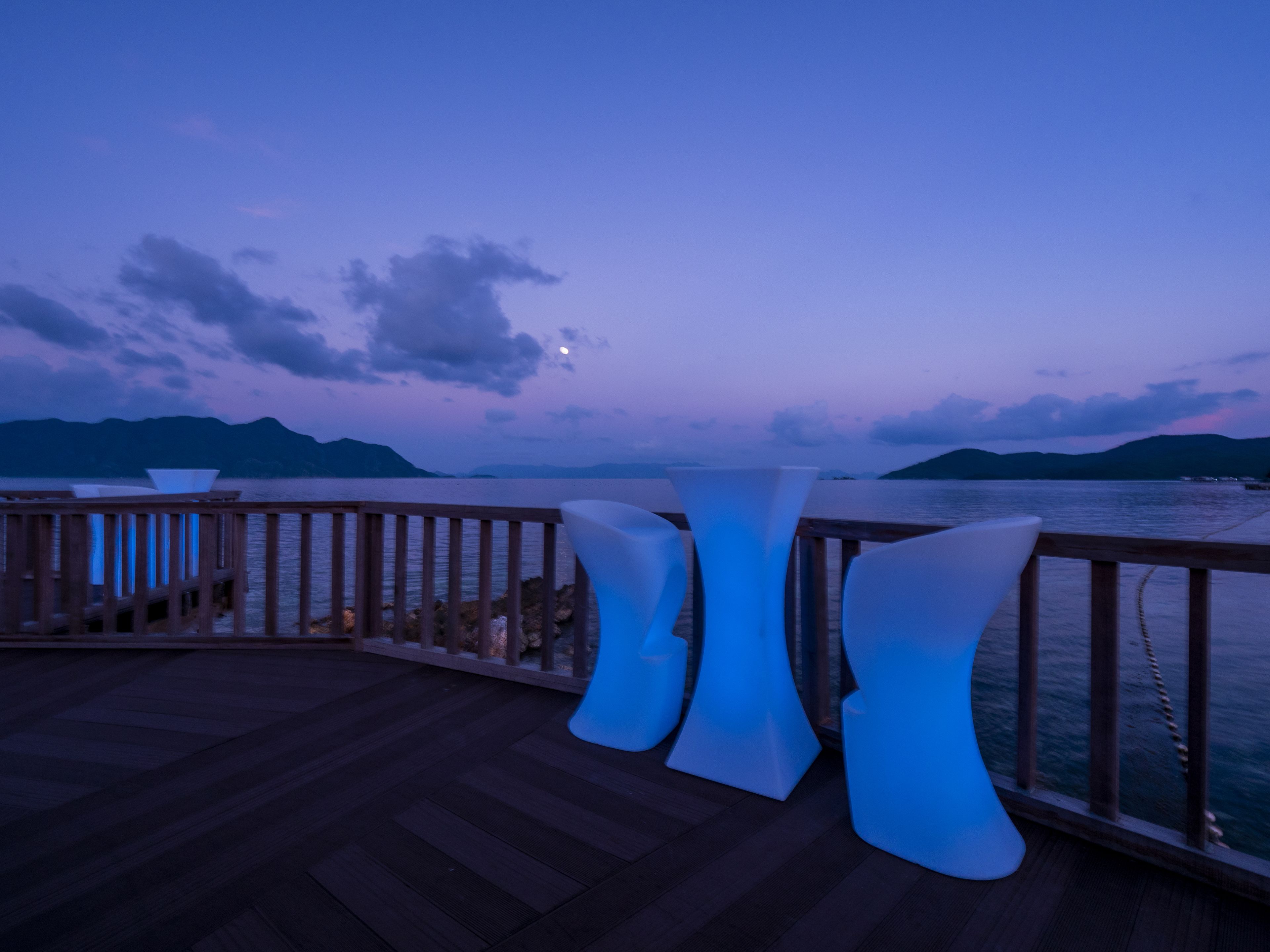 A deck with modern chairs and a table, overlooking a body of water and distant mountains.