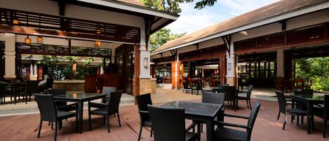 An outdoor patio area with tables and chairs, covered by a roof, and surrounded by greenery.