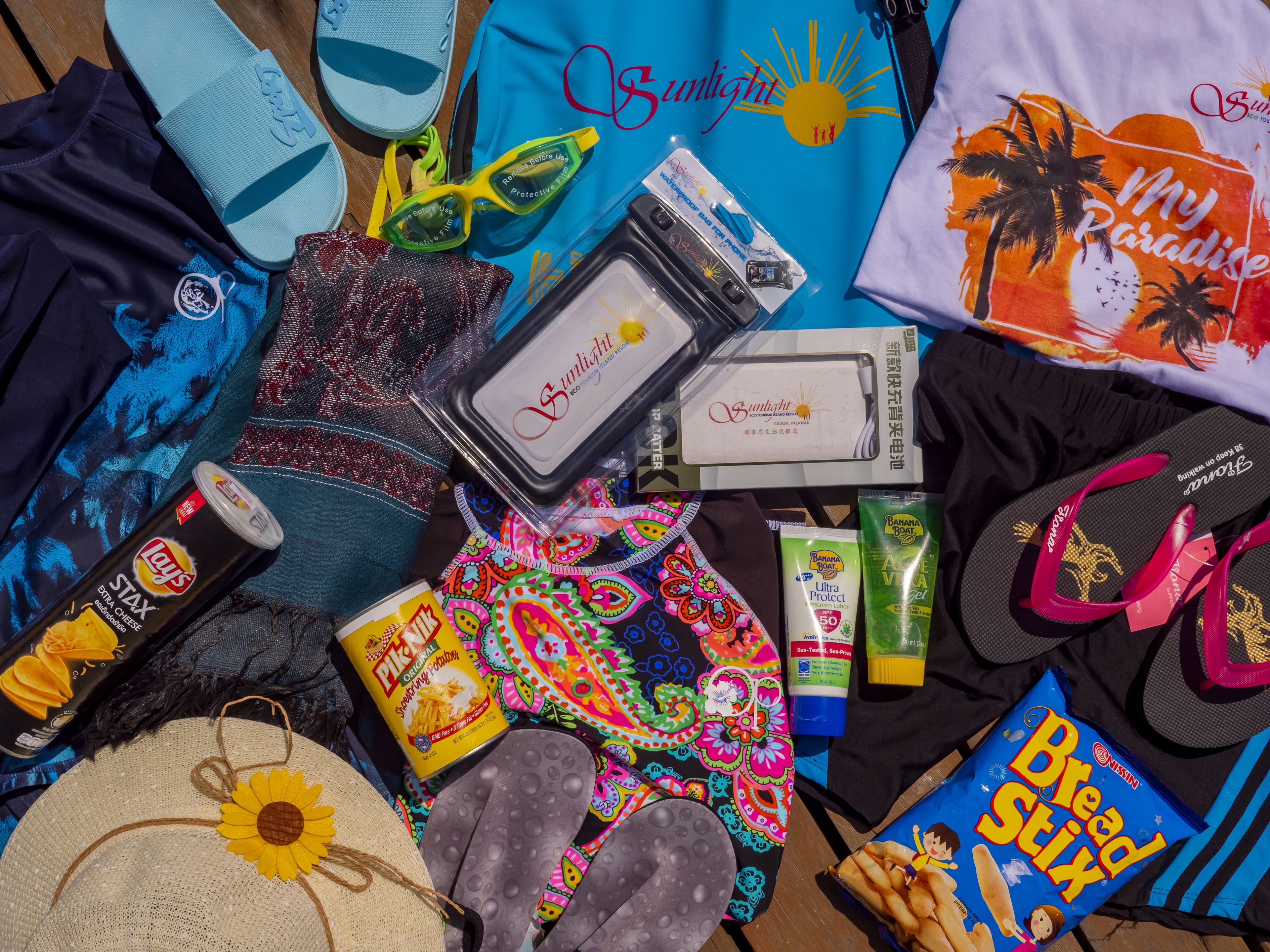 Diversos artículos de viaje como un sombrero de paja, chanclas, gafas de sol, una botella de protector solar, una botella de agua, aperitivos y una camiseta.