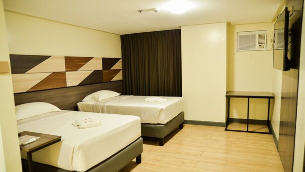 A hotel room with two beds, a small table, a television, and a wall-mounted air conditioning unit. - Travelite Hotel Legarda (Baguio)