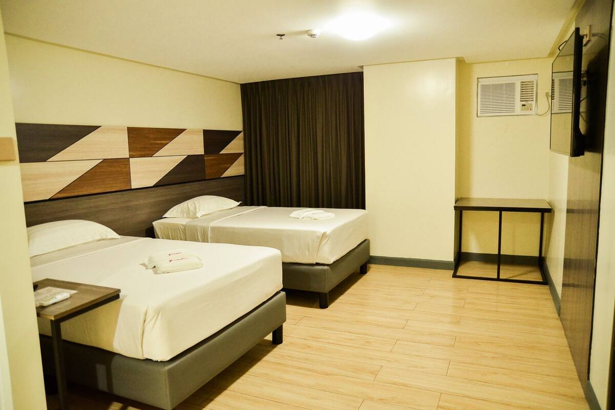 A hotel room with two beds, a small table, a television, and a wall-mounted air conditioning unit.