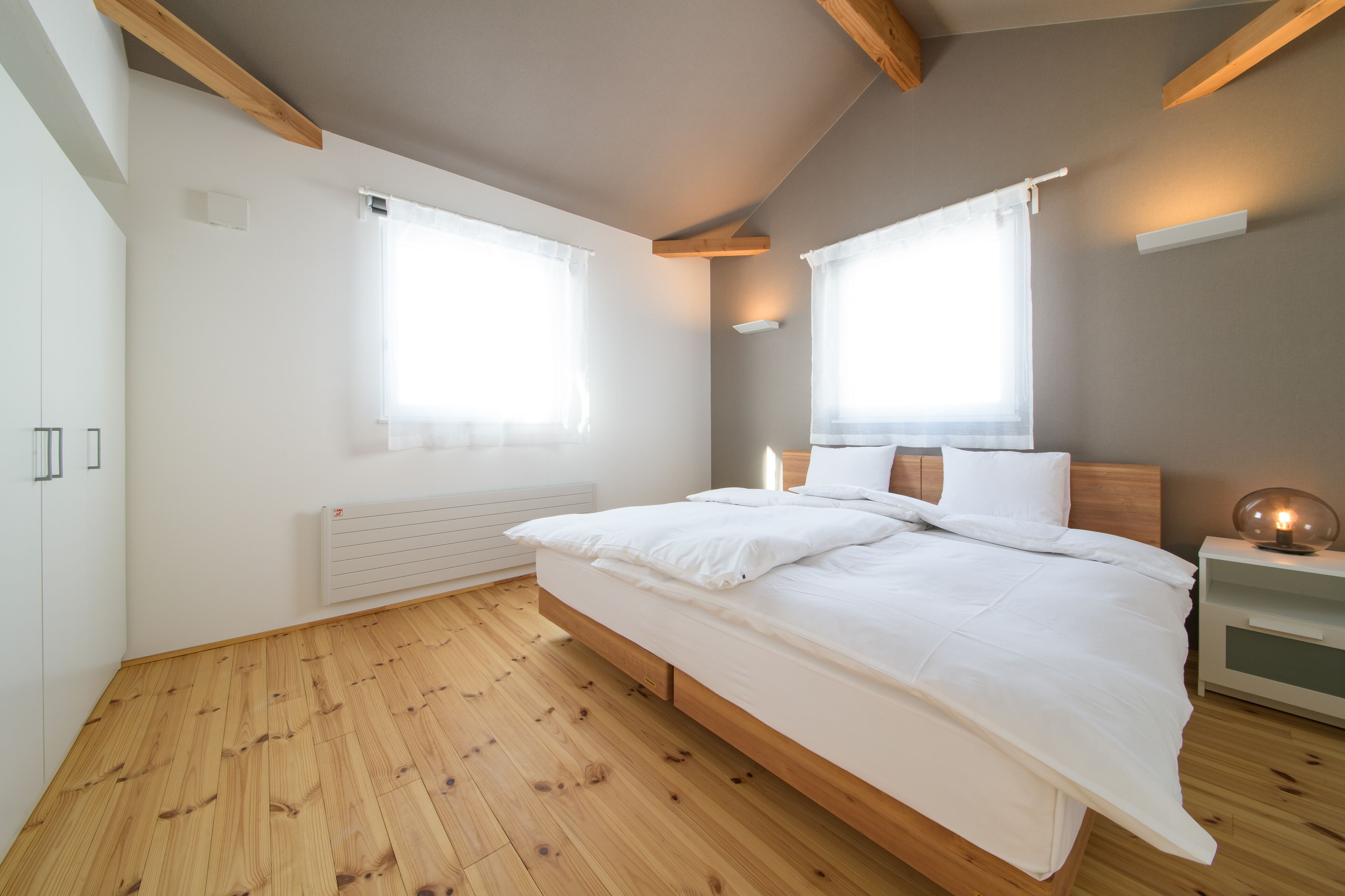 A bedroom with a wooden floor, a bed with white bedding, a bedside table with a lamp, and two windows with curtains.