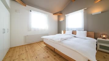 A bedroom with a wooden floor, a bed with white bedding, a bedside table with a lamp, and two windows with curtains.