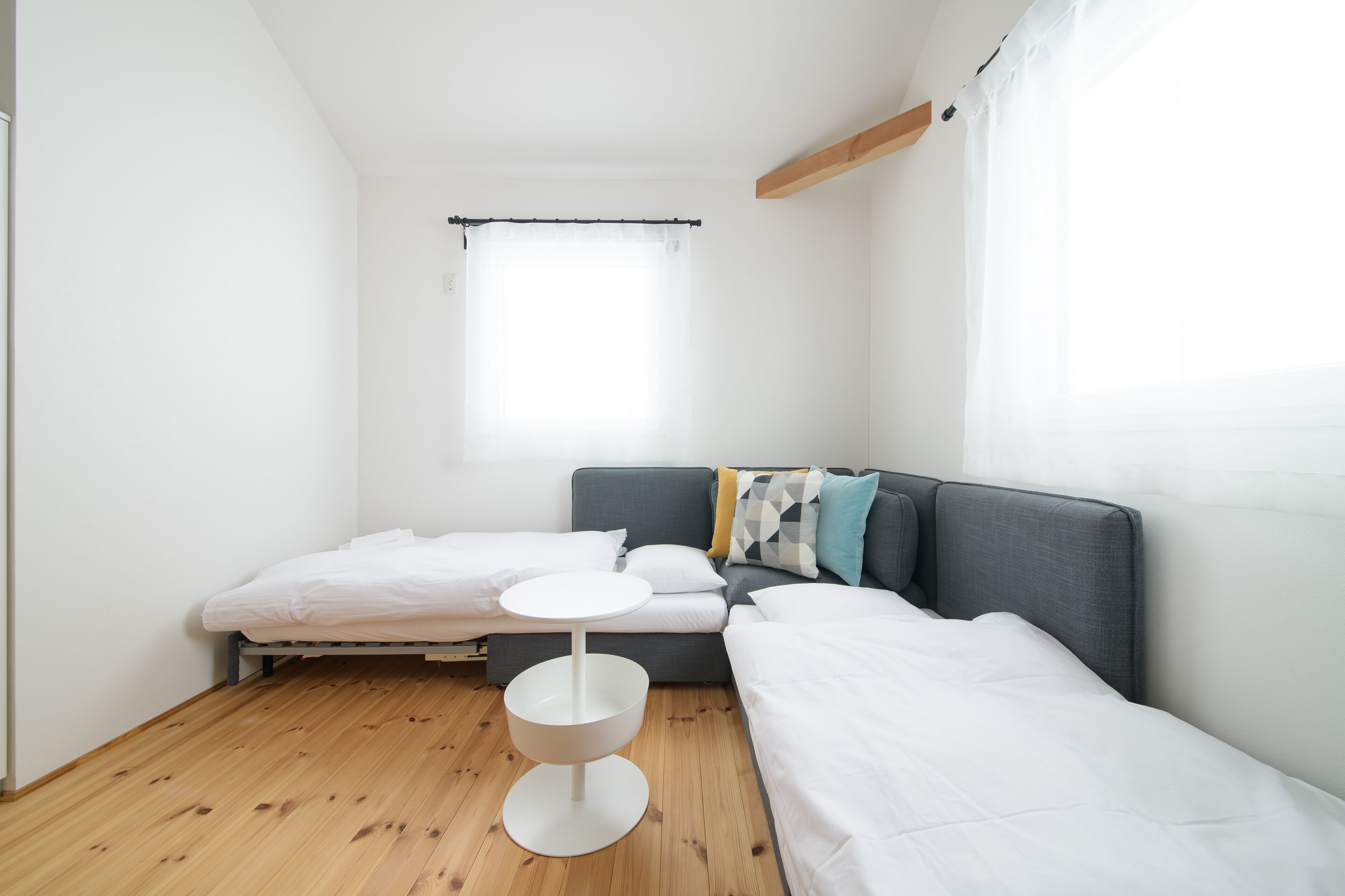 A modern, minimalistic bedroom with a grey sofa, two single beds, a small round table, and a large window with sheer curtains.