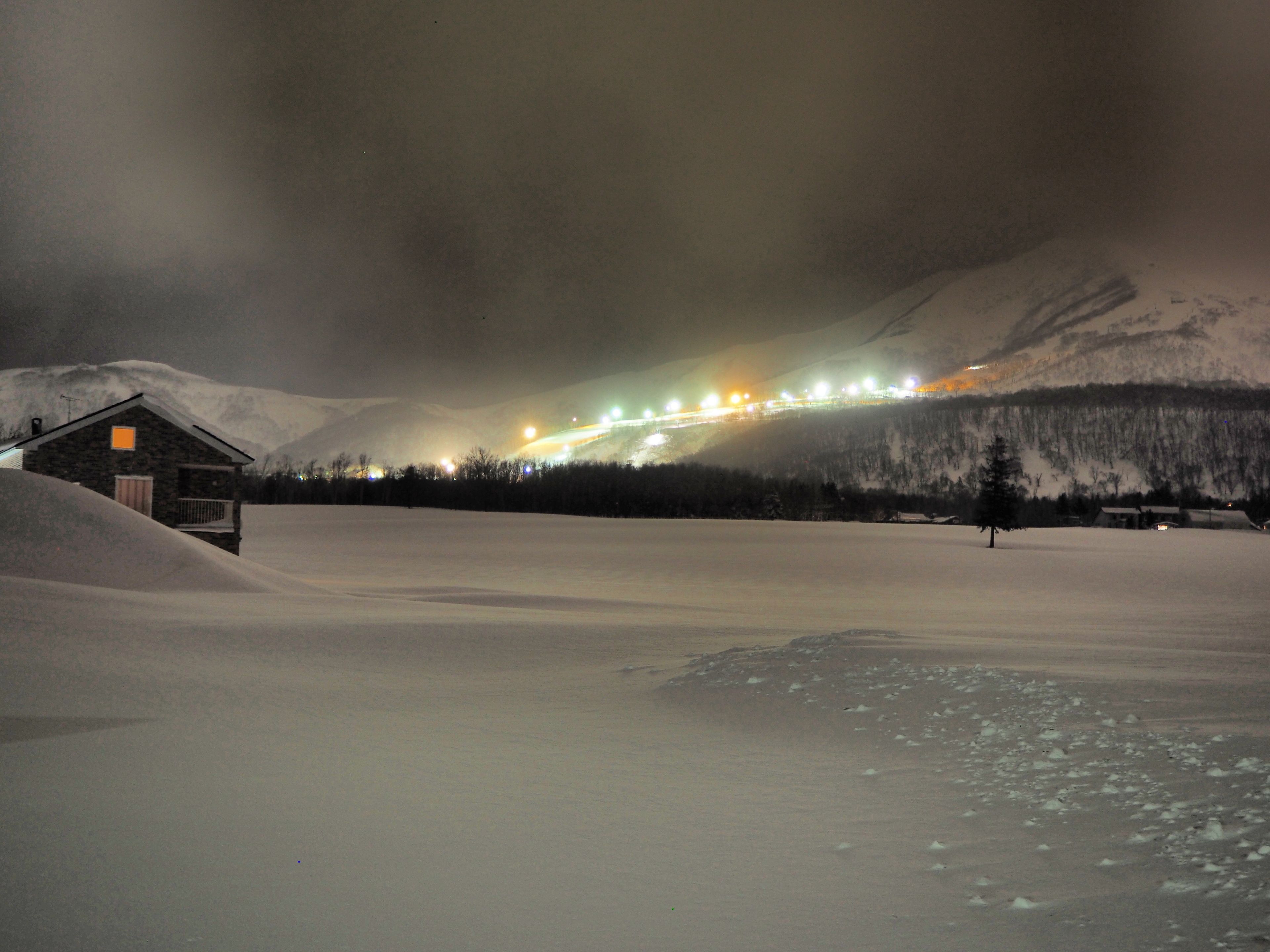 Paesaggio innevato con una piccola baita e una struttura illuminata in lontananza, probabilmente un treno o una strada, sullo sfondo di montagne.