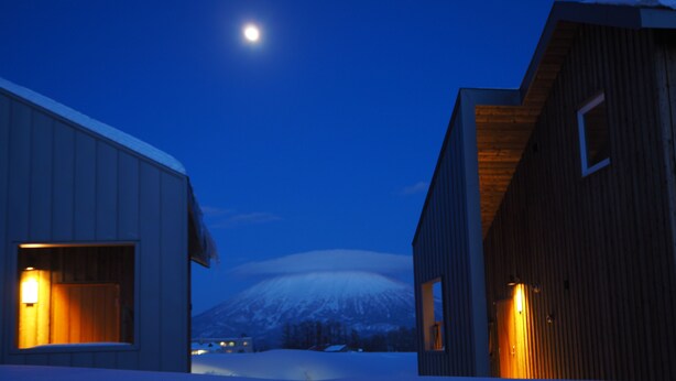 A snow-covered landscape with two buildings, one with a wooden exterior and the other with a white exterior, both with lit windows. - Niseko Highland Cottages (Niseko)