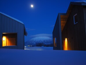 A snow-covered landscape with two buildings, one with a wooden exterior and the other with a white exterior, both with lit windows. - Niseko Highland Cottages (Niseko)