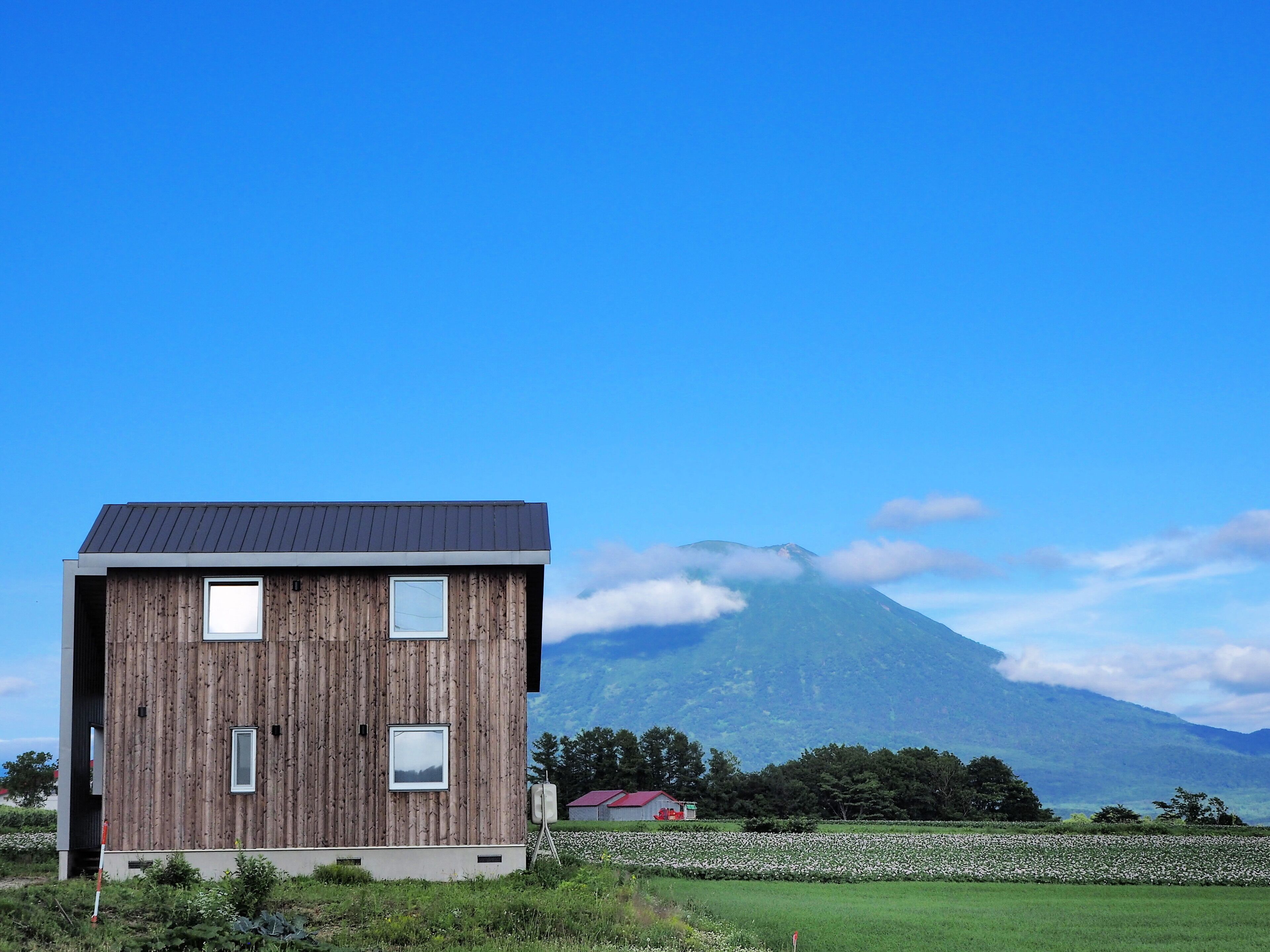 Une maison moderne en bois, dotée d’un toit plat et de grandes fenêtres, qui se détache sur un paysage de montagne et d’un ciel bleu azur.