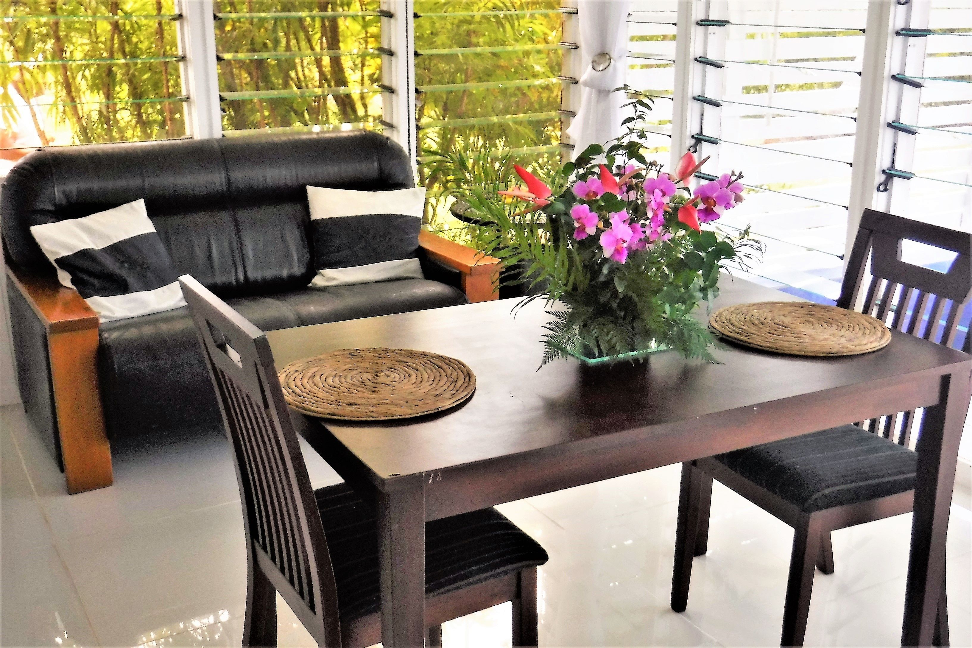 Ein Essbereich mit einem dunklen Holztisch, schwarzen gepolsterten Stühlen und einer schwarzen Ledersofa mit weißen Kissen. Auf dem Tisch liegt eine gewebte Tischmatte, und in der Tischmitte steht ein Blumentopf mit rosa Blüten.