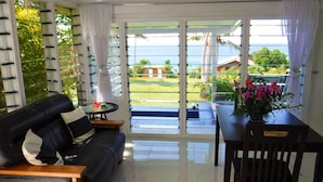 A living room with a black leather sofa, a wooden coffee table, and a dining table with a chair. There is a view of a balcony with a glass door leading to a garden and a view of the sea. - Makaira Resort (Taveuni Island East)
