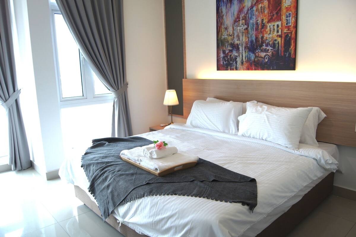 A neatly made bed with white linens, a grey throw blanket, and a tray with folded towels and a rose.