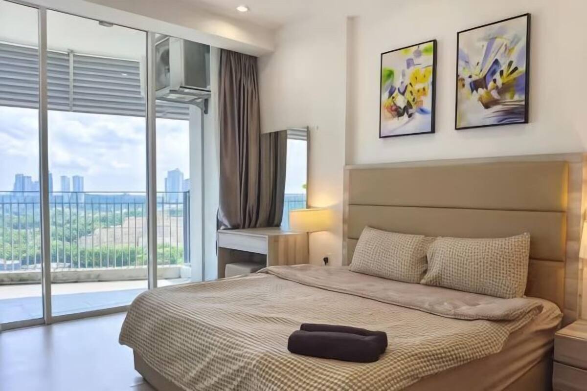 Un dormitorio con una cama grande, una mesita de noche, un escritorio y vistas a un paisaje urbano a través de una puerta corrediza.