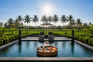 Una piscina con una bandeja flotante de alimentos para el desayuno, rodeada de palmeras y un cielo despejado.