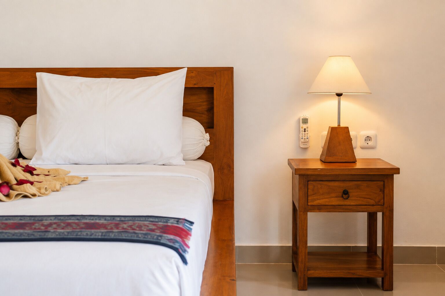 Una cama bien hecha con sábanas blancas y una sola almohada, una mesita de noche con una lámpara y un termostato fijado en la pared.