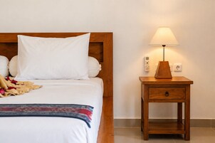 Cama bem arrumada com lençóis brancos e um único travesseiro, mesa de cabeceira com abajur e termostato fixado na parede.