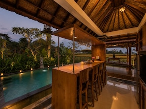 Zona de bar con vistas a la piscina, muebles de madera y techo de paja.