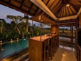 Zona de bar con vistas a la piscina, muebles de madera y techo de paja.