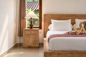 A hotel room with a wooden headboard, a bedside table with a lamp, a bed with white bedding, and a window with curtains. - Griya Private Villa (Ubud)