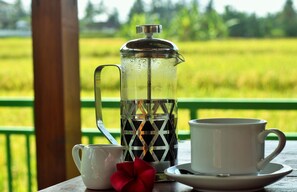 A clear glass French press with a metal handle and a lid, placed on a wooden table with a white cup and saucer beside it. - Griya Private Villa (Ubud)