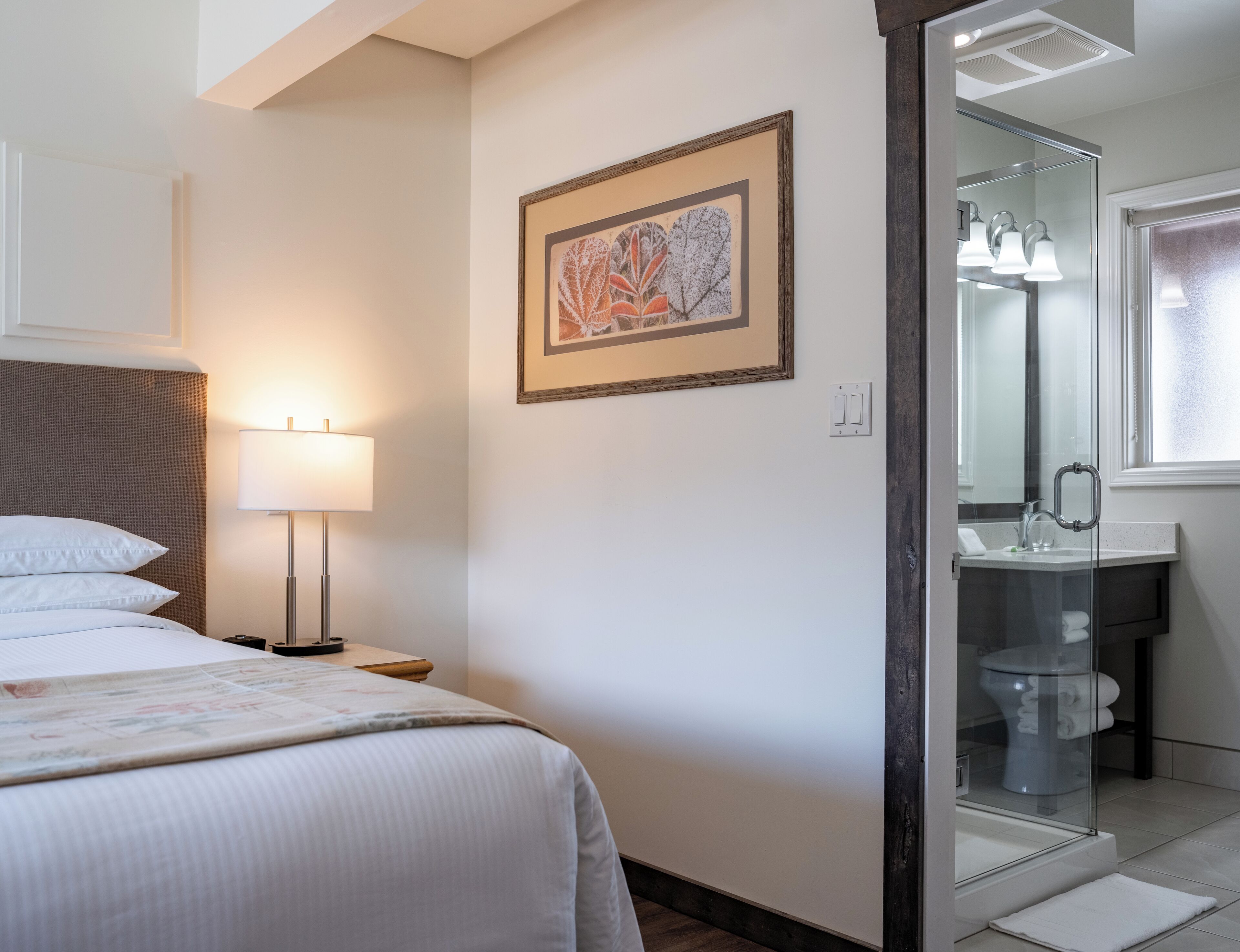 Habitación de hotel con cama, lámpara de noche, cuadro enmarcado y baño con cabina de ducha de vidrio.