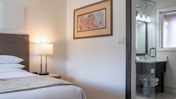 Et hotelværelse med seng, sengebordlampe, indrammet kunst og et badeværelse med glasbrusekabine.