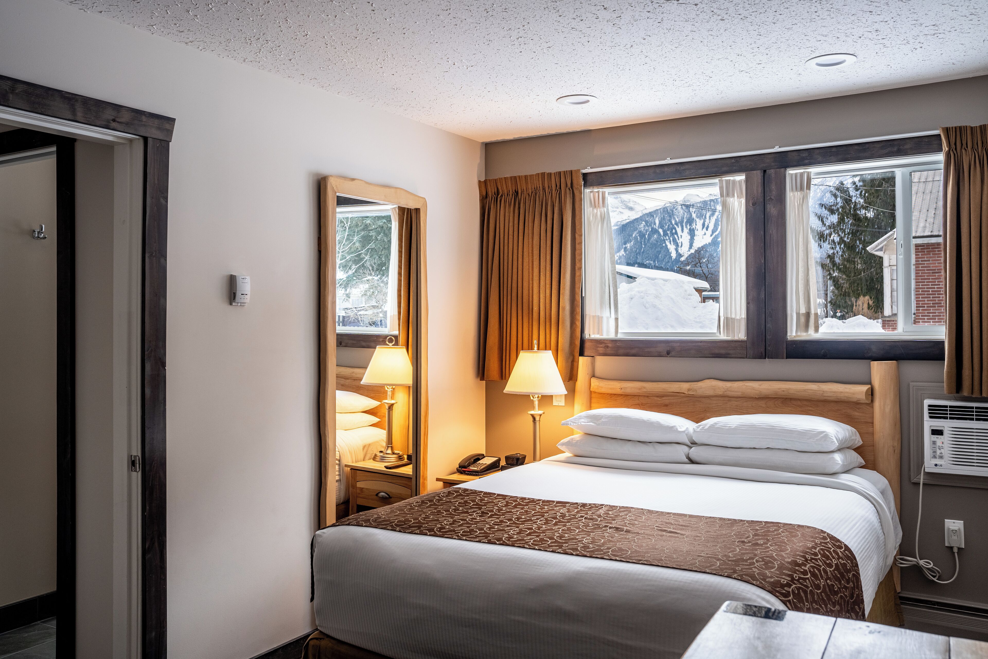 Une chambre d’hôtel avec un grand lit, deux lampes de chevet, une fenêtre avec des rideaux, un miroir et un thermostat.