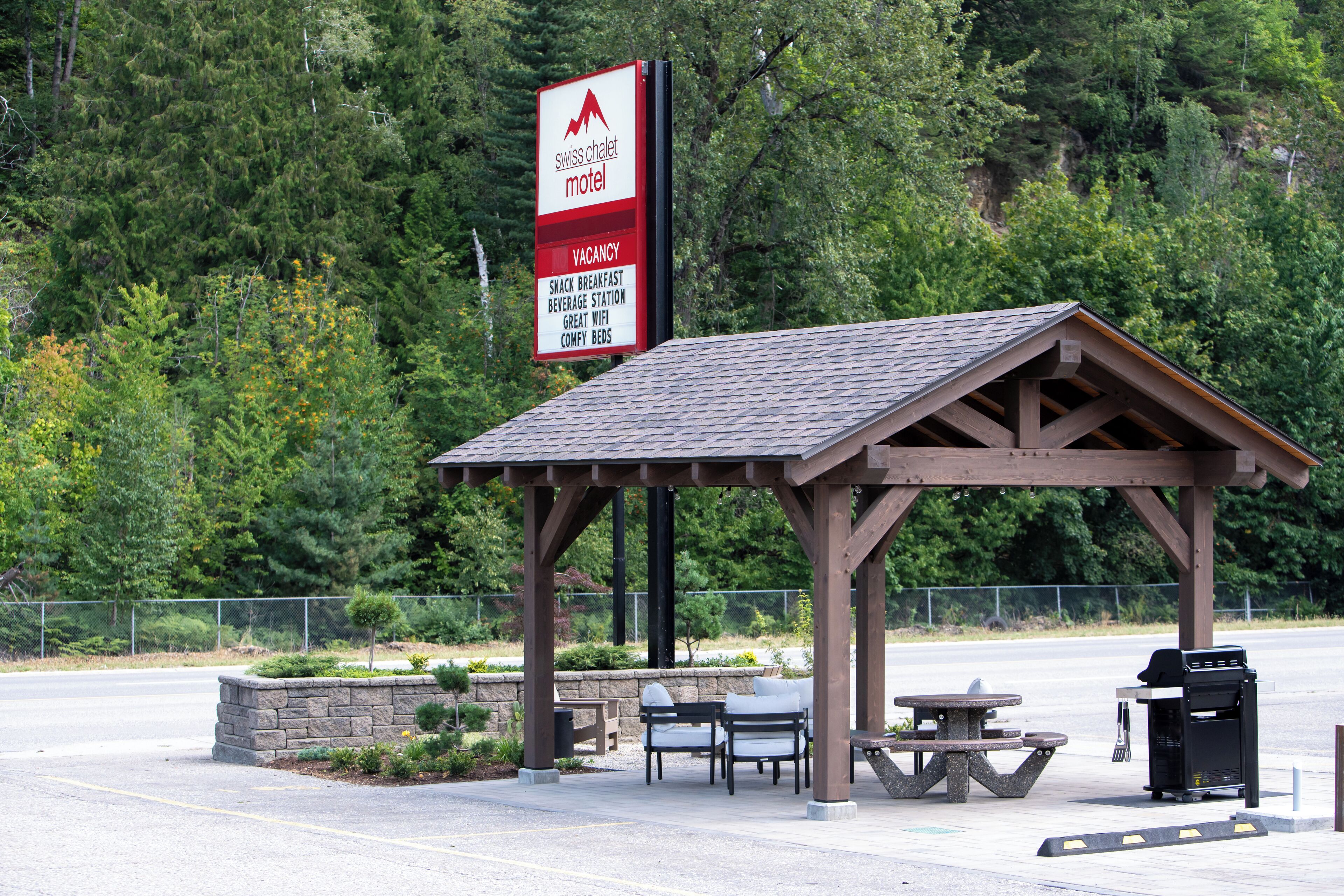 A Swiss Chalet Motel sign with amenities listed, a covered picnic area with tables and chairs, and a stone planter with plants.
