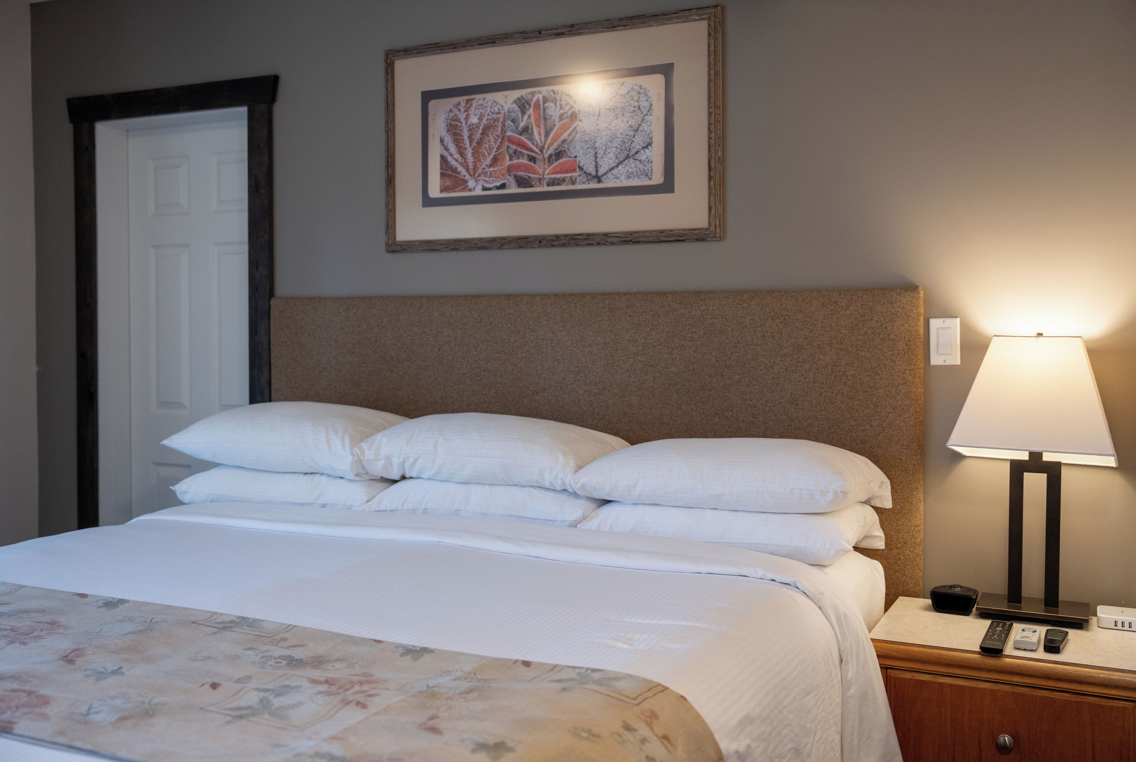 A neatly made bed with white linens and a floral-patterned bedspread, a headboard, a bedside lamp, and a framed artwork on the wall.