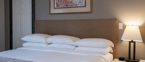 Una cama bien tendida con sábanas blancas y una colcha de estampado floral, un cabecero, una lámpara de noche y una obra de arte enmarcada en la pared.