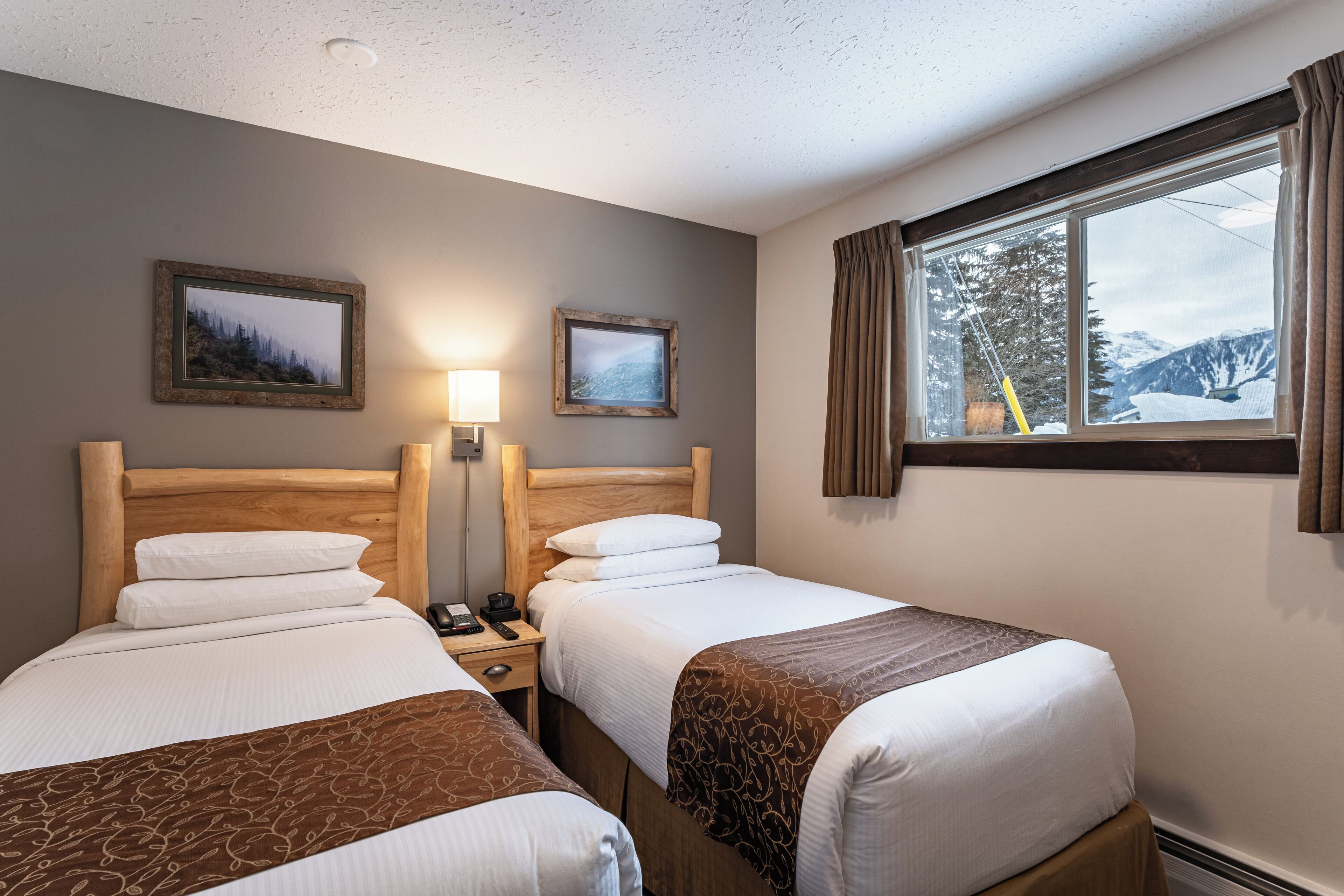 Ein Hotelzimmer mit zwei Betten, Holz-Kopfteilen, einem Nachttisch mit Lampe und einem Fenster mit Blick auf schneebedeckte Berge.
