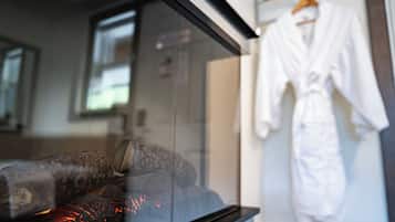 A glass partition with a visible fireplace, a white robe hanging on a rack, and a window with blinds.