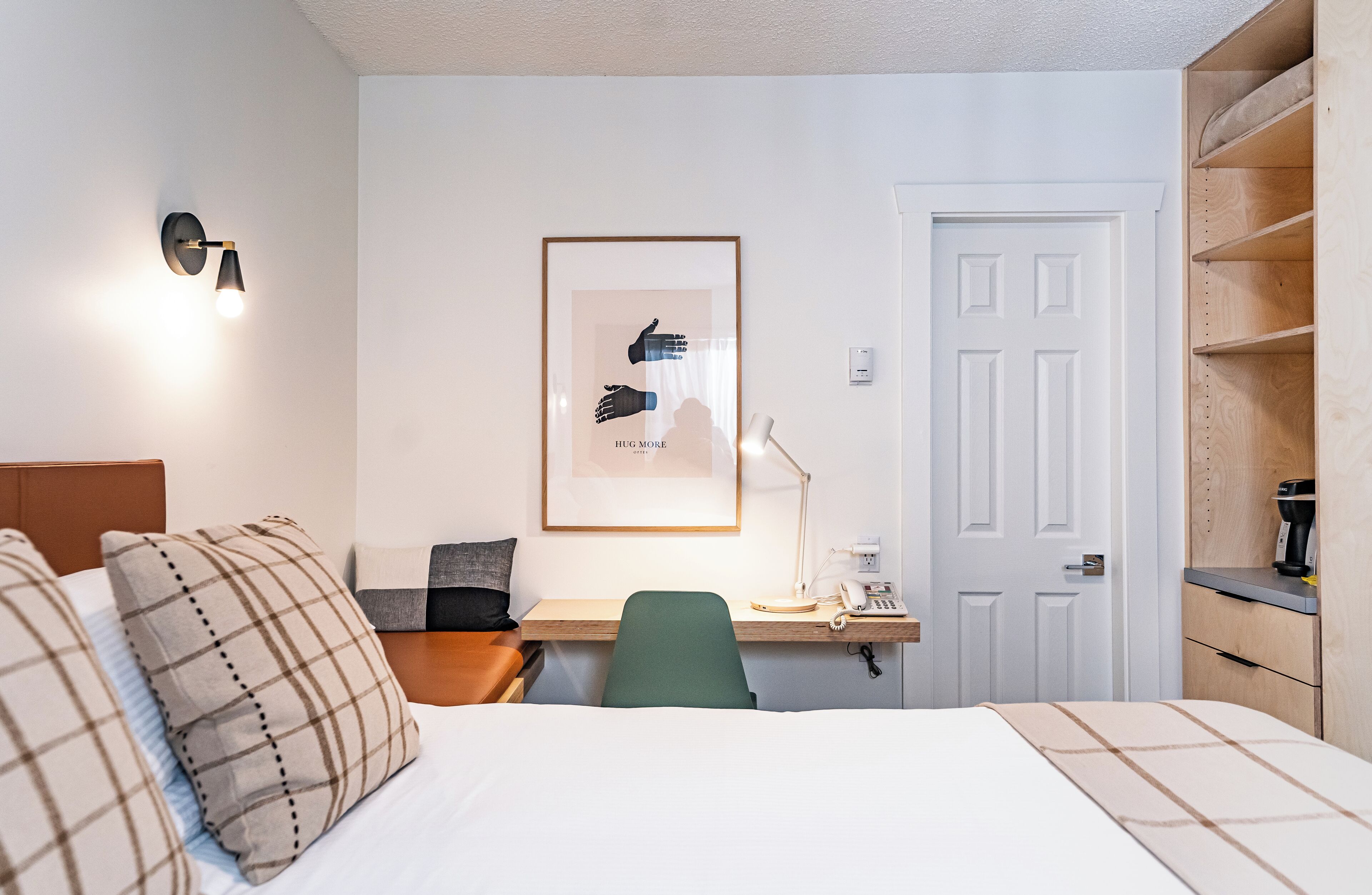 A hotel room with a bed, a desk with a chair, a framed picture on the wall, a door, and a shelf with a coffee maker.