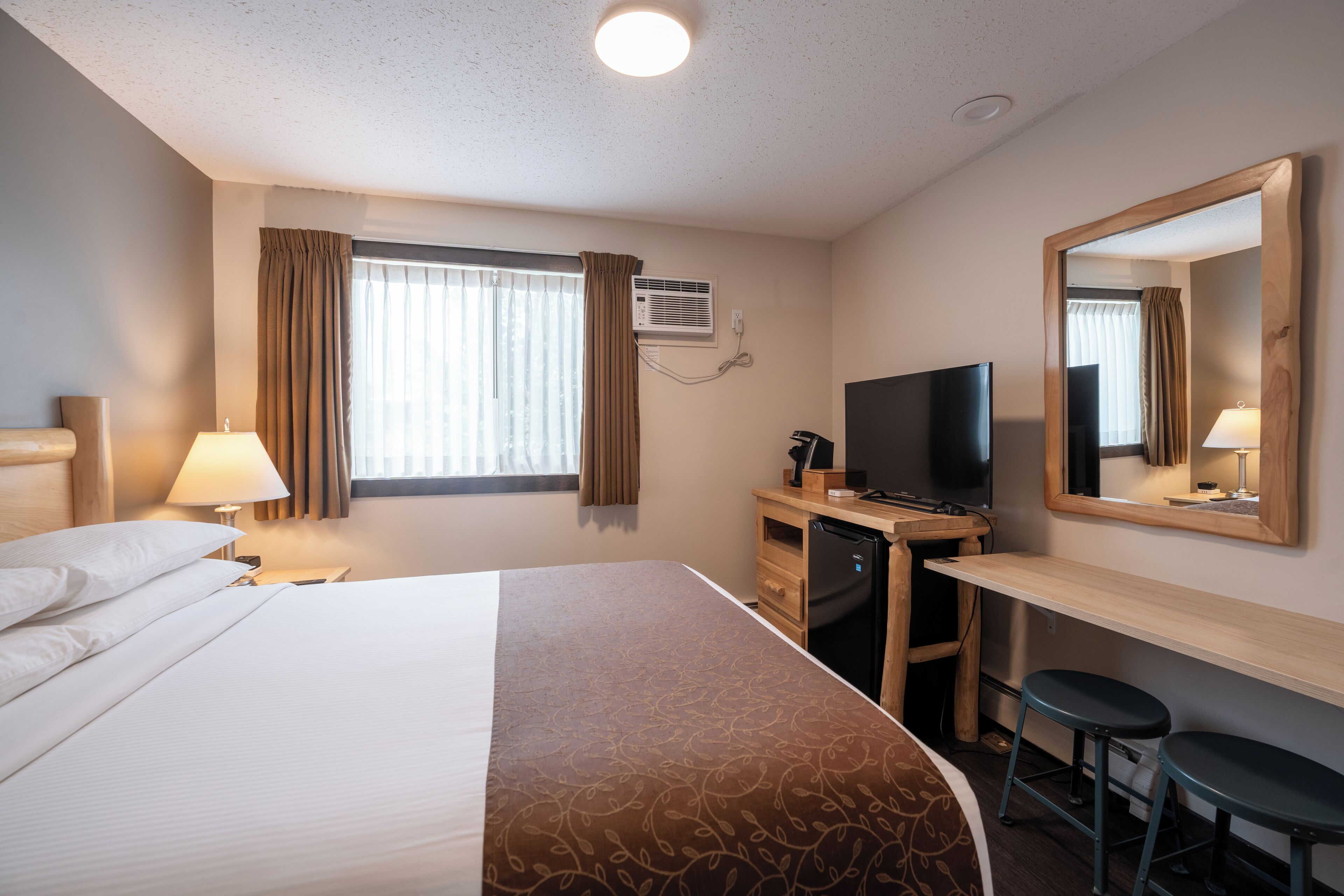 A hotel room with a bed, desk, TV, and two stools.