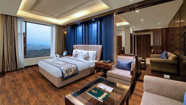 Una habitación de hotel moderna con una cama grande, una zona de estar con sofá y sillón, una mesa de centro de vidrio y vistas a las montañas.