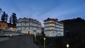 Un hotel de varios pisos con una fila de coches aparcados en la parte delantera, ubicado en una colina bajo un cielo despejado.